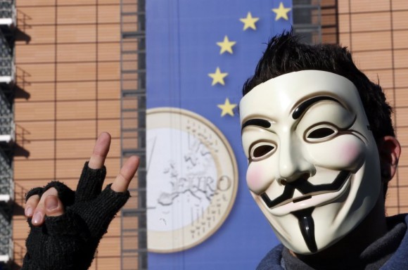 Un joven con una máscara de Anonymous frente a la Comisión Europea, en Bruselas (Bélgica), con la moneda de euro en su fachada. Foto: Reuters.