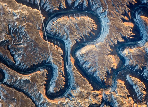 Este sector del cañón del Río Verde en el este de Utah se conoce como Curva del Bowknot debido a la forma en que el río se dobla sobre sí mismo. En esta fotografía tomada por un astronauta en la Estación Espacial Internacional el 22 de enero de 2014, el Green River aparece oscuro debido a que se encuentra en la sombra, 300 m (1.000 pies) por debajo del paisaje circundante. Los acantilados de color amarillo con tintes tamizados por la salida del sol dan una idea de las paredes empinadas del cañón. La línea blanca recta a través de la escena es la estela de un jet liner volando sobre el cañón. Foto: NASA. 